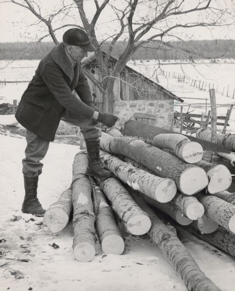 A man sawing the top log in a pile of firewood with a handsaw. There is snow on the ground, and he is dressed warmly. Behind him is a tree, a stone and wood building. Fields and a forest are in the distance. 