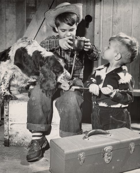 Two young boys are oiling the reel of a fishing rod in a shed. The older boy is sitting on a box, and the younger boy is kneeling. A Springer Spaniel has his front feet on the box and is sniffing the oil. A toolbox is in the foreground.