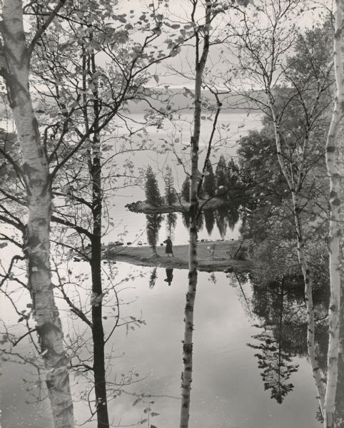 Elevated view towards a woman posing on a spit of land jutting into a lake, seen through birch trees in the spring. Caption reads: "Spring comes suddenly to Wisconsin and brings a wondrous world of sunny skies, fresh green fields and budding trees. There's a mighty vigor in the air that sets blood racing, footsteps moving faster."
