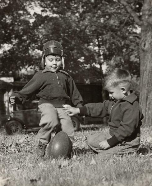 Two young boys playing with a football. One boy wearing a helmet is about to kick the football, just as the other boy has just stood the ball upright.