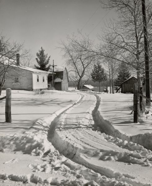 Tire tracks in fresh snow lead into a barnyard with a farmhouse and farm buildings.