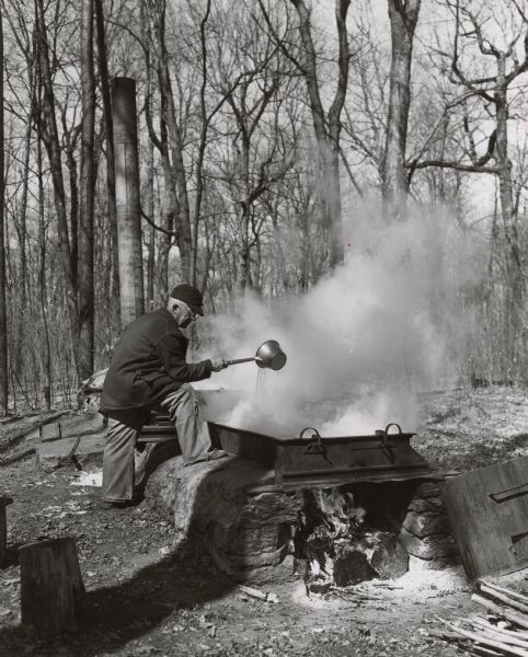 A man is cooking maple syrup over a wood fire among trees. Steam is billowing from the kettle.