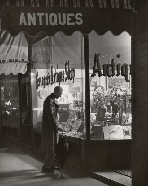 A man carrying an umbrella is gazing into the display window of Levinger's Antique Shop, Jefferson and East Main Street.