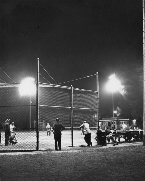A tense moment in a night baseball game played at the Dorothy Enderis Playground. Two men are standing and watching the game through the chain link fence.
