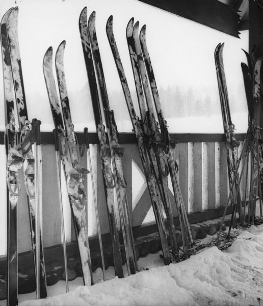 Snow covered skis are lined up against the porch railing while their owners are taking a break in the ski lodge. Pine trees can barely be seen in the distance through the light snow.
