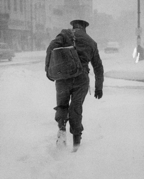 A mailman is trudging through the snow with his loaded mailbag. A city street and buildings are barely visible through the snow.