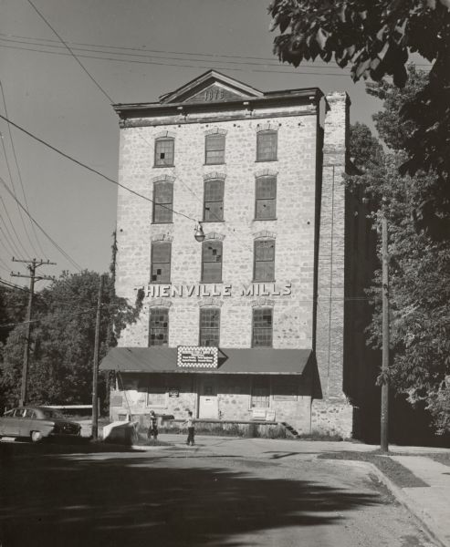 View down road towards two people walking in front of the Thienville Mills, a five-story stone building, built in 1876. At this time, it was the Thiensville Feed and Supply Company. Trees are on the right and an automobile is on the left.