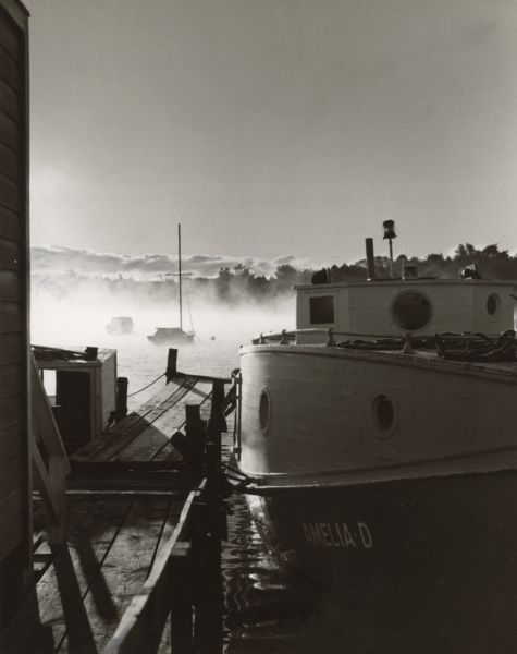 The <i>Amelia-D</i> is docked at a pier on Green Bay. The shoreline and clouds are in the background. A sailboat and motorboat are moored in the distance.