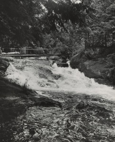 Water cascades beneath a footbridge and over boulders on a river. A canopy of trees is overhead.