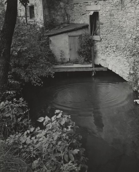 Pool at an old distillery in Hamilton. On the left are trees and shrubs. On the right is a stone building that is arched over the pool. Above the arch is a bricked up window with a sign attached the lintel that reads: "Warning, Keep Away." To the left below the window is a small roofed structure with a latched door that is sitting on a wooden pier just above the water.