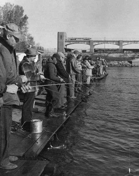 View of a crowd of people lined up fishing side by side on long pier. A railroad bridge, with a tanker car, is in the background.