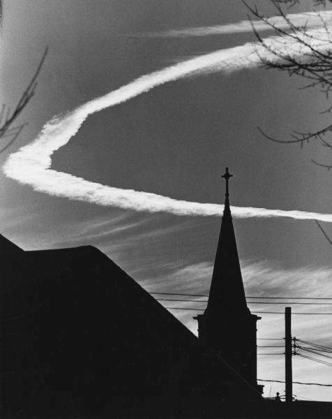 A circular contrail forming a halo behind a silhouetted church steeple. Power lines are in the background.