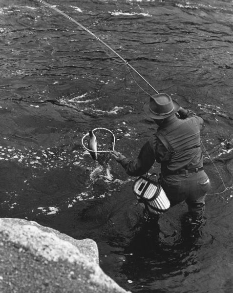 Elevated view of a fisherman netting his fish near the bridge at Keshena Falls. Caption on reverse reads: "This is an actual catch I just happened to be present with my camera as this man caught a trout."