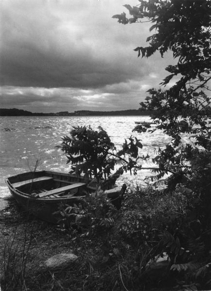 View from shoreline towards a rowboat is pulled up onto the shore of Squaw Bay on Lake Monona. Trees are in the foreground. The sky is overcast and the water is choppy. The far shoreline is in the background.