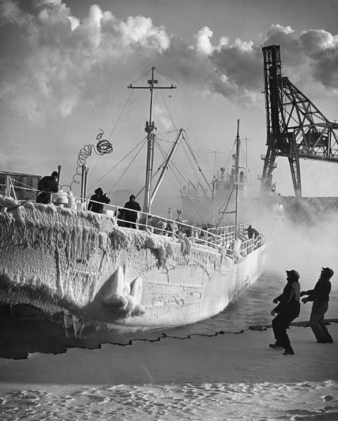 A man on a ship is tossing a coiled rope to two men below on the wharf. Ice encrusts the ship. Several other men are standing on the deck. Another ship and a gantry are in the background.
