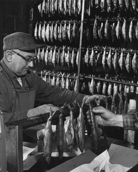 Two men are packing smoked fish in a box. In the background smoked fish are hanging on nails on racks.