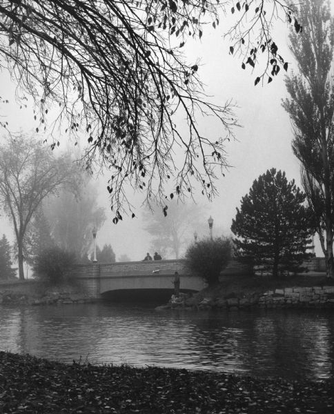 View across water towards two men standing on a stone bridge, in the early morning fog in September. The bridge is spanning one of the canals. Another man is standing on the shoreline below them, fishing from the rocks.
