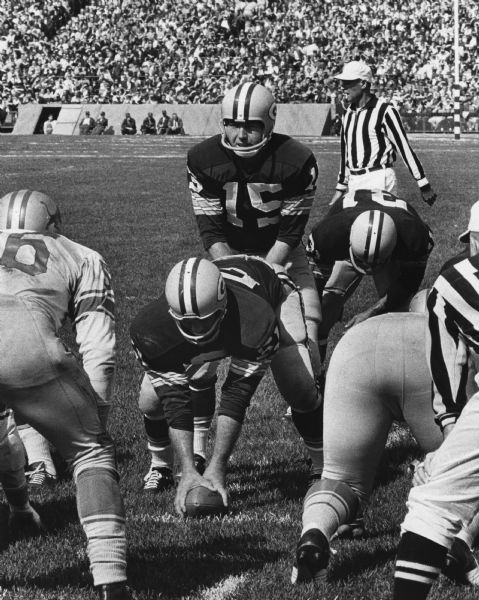 Green Bay Packer's Quarterback Bart Starr and Center Jim Ringo in formation during a football game against the Detroit Tigers. Two referees observing, and spectators are in the stands in the background.