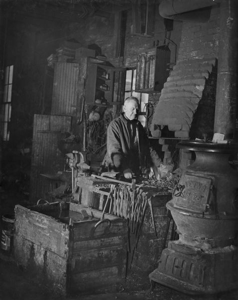 A man at the forge in Albert Smith's blacksmith shop. On the right is an Aetna No. 25 stove. Tools are hanging on the edge of the forge.