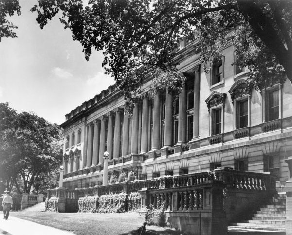 View of the front of the Wisconsin Historical Society. A man is walking down the sidewalk on the left. Vines cover the stone balustrade. A tree is in the foreground on the right.