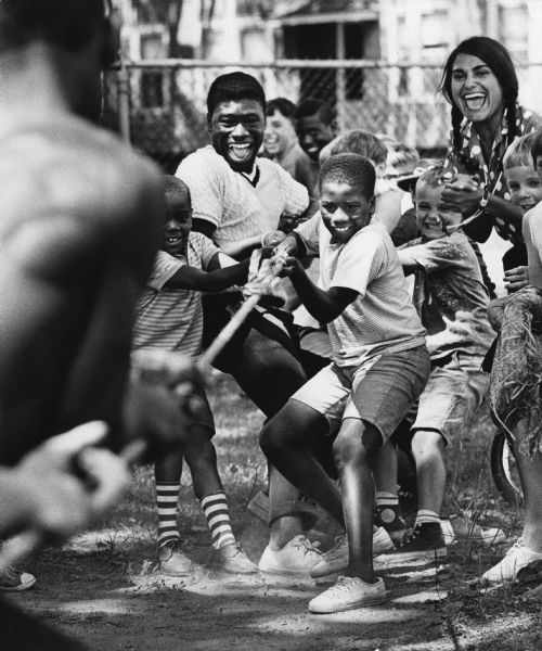 Children and adults having a tug of war during a play day at Marquette School. A fence and dwellings are in the background.