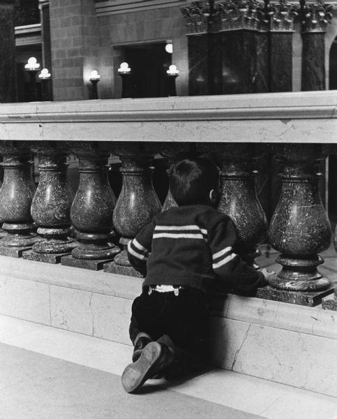 A young boy is kneeling and peering in between balusters of a balustrade indoors at the Wisconsin State Capitol.