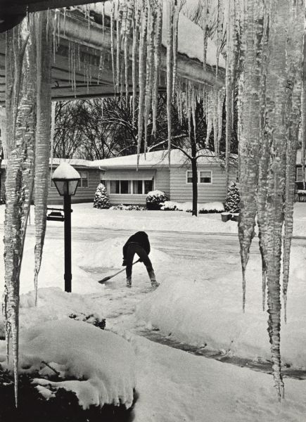 A man shoveling his driveway. He is framed by large icicles hanging from the eaves of the house. Across the street are more homes, and trees.