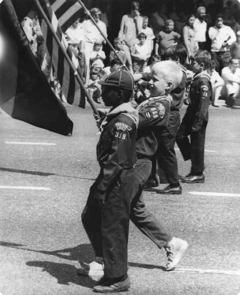 Cub Scouts marching in a Memorial Day parade. Two scouts are holding flags, and spectators are in the background.