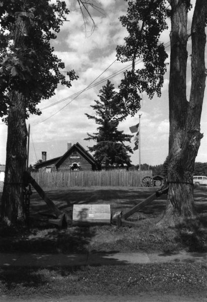 View of the Madeline Island Historical Museum and a fence framed by two trees. A wooden sign points the way.