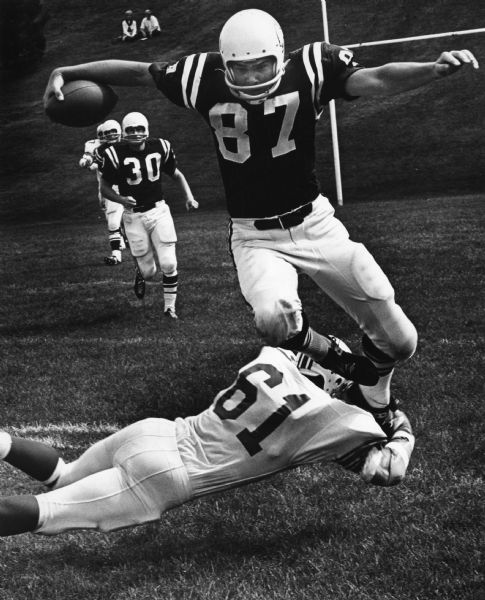 A football player is leaping to avoid a tackle at Lawrence University. Two people are watching from the hillside in the background.