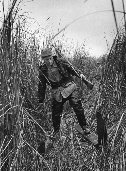 A duck hunter with his gun on his shoulder navigates a marsh using swamp skis. Another hunter is standing in the background.