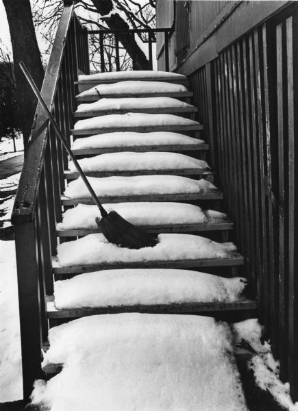 New snow on the steps at Devil's Lake, with a broom leaning on the hand rail. Trees are in the background.