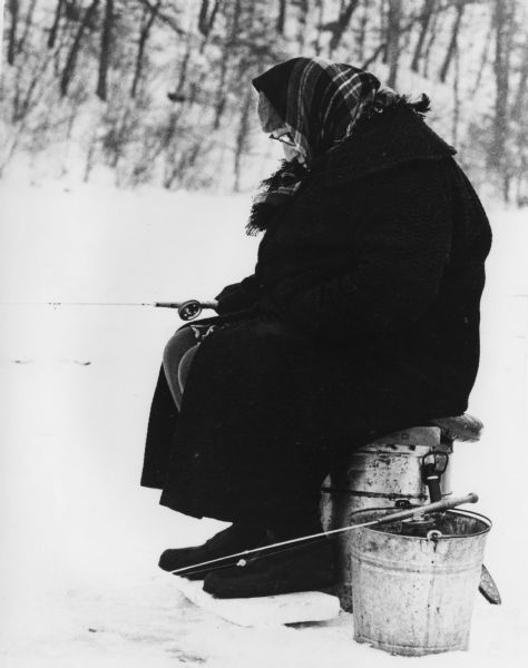 A woman wearing a hat and a plaid scarf over her head is ice fishing in Governor Dodge State Park. She is sitting on a bucket with her feet on a board.