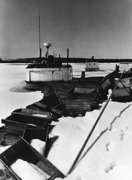 Fishing boats wintering on Lake Superior, which is frozen and covered with snow. Wooden fishing boxes are strewn in the foreground, and a wooded shoreline is in the distance.