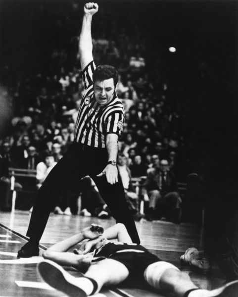 A referee has his fist raised over his head indicating a foul while standing over a basketball player lying on the floor. They are at the State Boys High School Basketball Tournament.