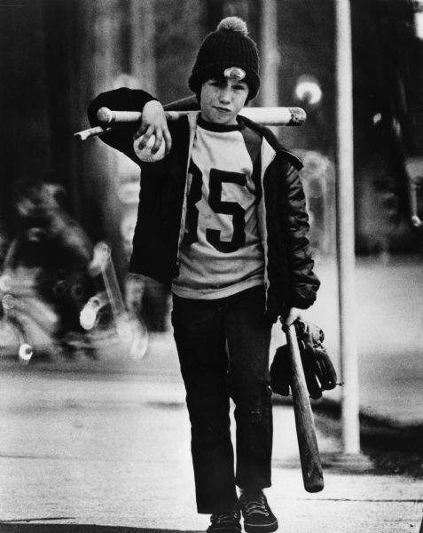 A boy is walking towards the camera carrying three bats, a baseball mitt and a baseball. His shirt displays the number 35 or 85.