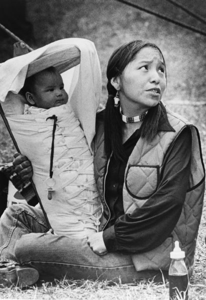 A young native American woman is sitting on the ground holding her infant in a cradleboard on her lap, a traditional way to protect and transport babies. She is wearing traditional jewelry.