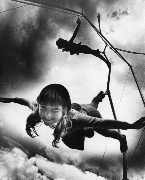 A girl tries some acrobatic moves on a rope at Acrobat School, Circus World Museum, a Wisconsin Historical Society site. The silhouette of another acrobat is above her on the rope. Both children have one foot in a loop on a rope.