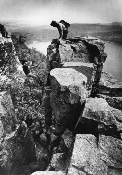 View towards a rock climber on top of an outcropping at Devil's Lake State Park who is watching another climber descending. Devil's Lake and bluffs are in the background.
