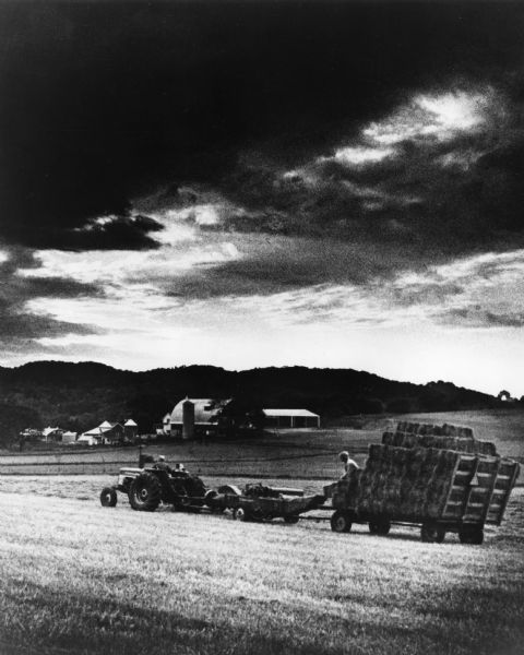 Farmers head for the barn with a load of hay bales as a storm looms overhead. A hay baler is hitched between the tractor and the wagon. Farm buildings are in the background.