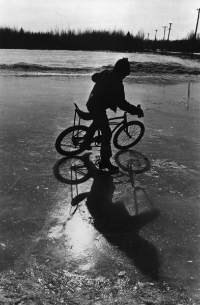 A boy is riding his bicycle on the ice. A field and a line of trees are along the shoreline in the background.