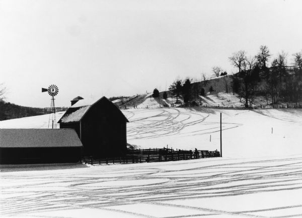 Snowmobile tracks form a pattern in the snow on a farm. Farm buildings are on the left and trees are along a ridge on the right.