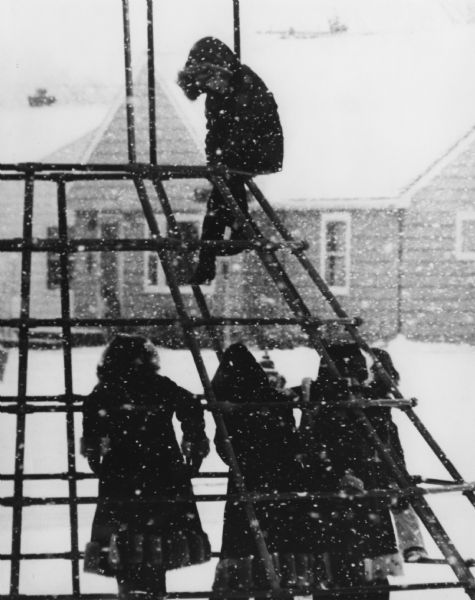 Children playing on monkey bars as snow falls. A dwelling or schoolhouse is in the background.