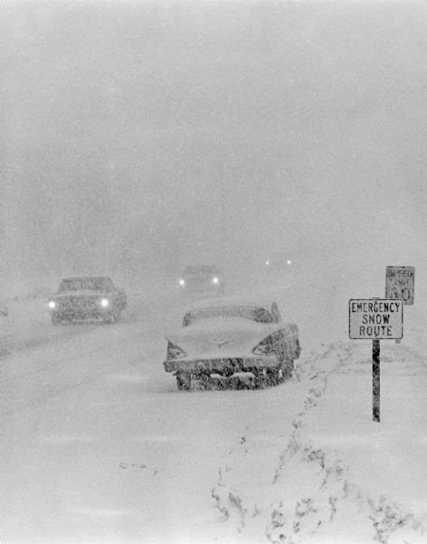 Automobiles are traveling through a winter storm on snow-covered roads.