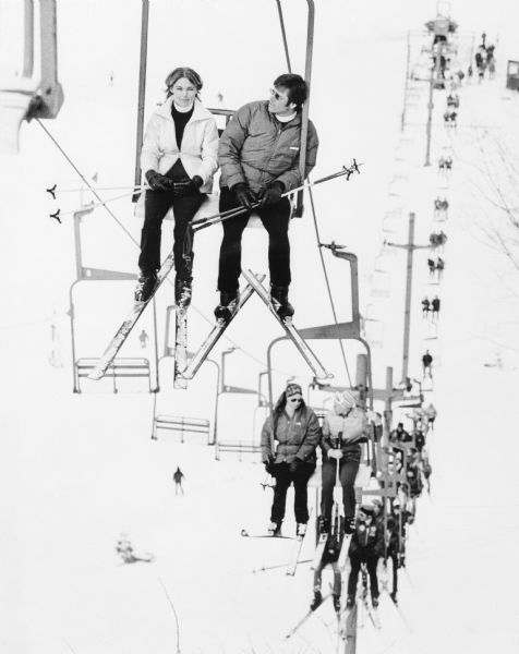 Elevated view of skiers gliding uphill on a chair lift at Wintergreen Ski Area. They are dressed warmly, and are wearing skis and carrying poles.