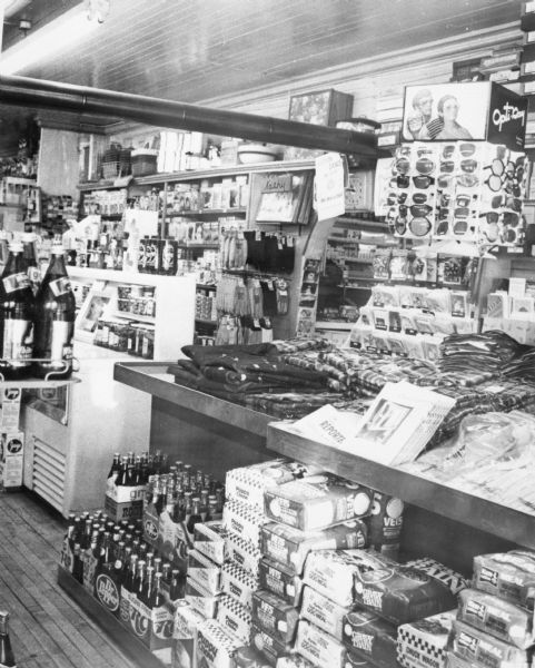 Interior view of a general store. Items are stacked on the floor, tables and shelves.