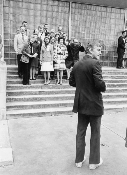 Classmates gathered outdoors during a reunion group portrait. They are posing on the steps of the Wisconsin Union Theater. One of the photographers in the foreground is looking off to the right.