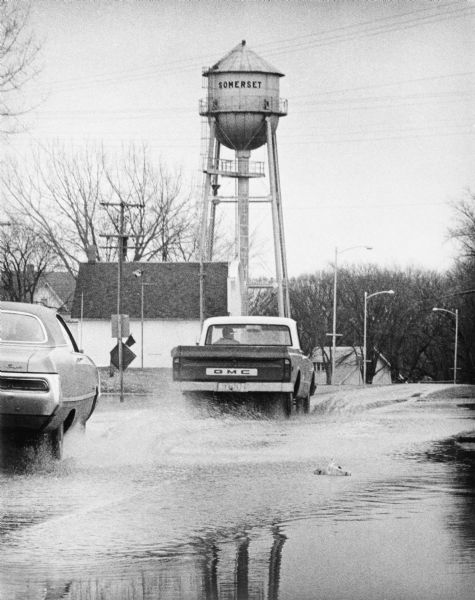 A GMC truck and an automobile are splashing through a large puddle in a small town. The town's water tower is in the background.