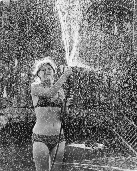 A woman spraying water upwards with a hose to create a "rain shower." She is wearing a bikini.