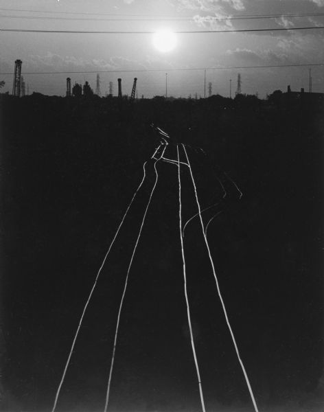 Elevated view of railroad tracks reflecting the light of the sun at either sunrise or sunset. Silhouettes of cranes, power lines and buildings are in the distance.